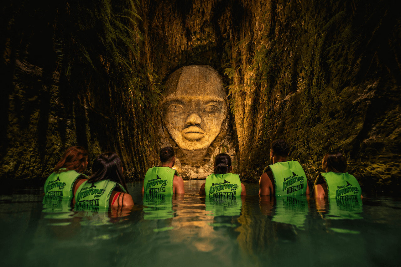 Visitantes con chalecos en el río subterráneo frente a una escultura de piedra iluminada