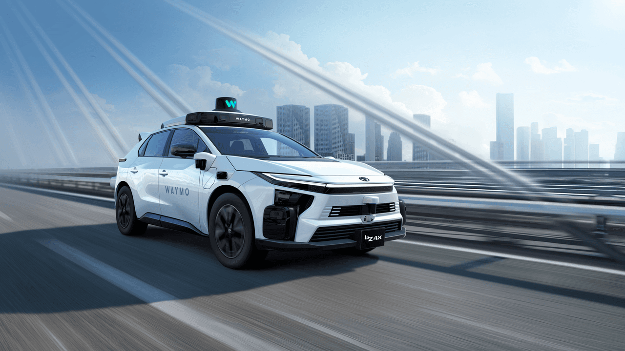 Toyota bZ4X blanco con el sensor de Waymo en el techo, rodando por un puente con rascacielos de fondo