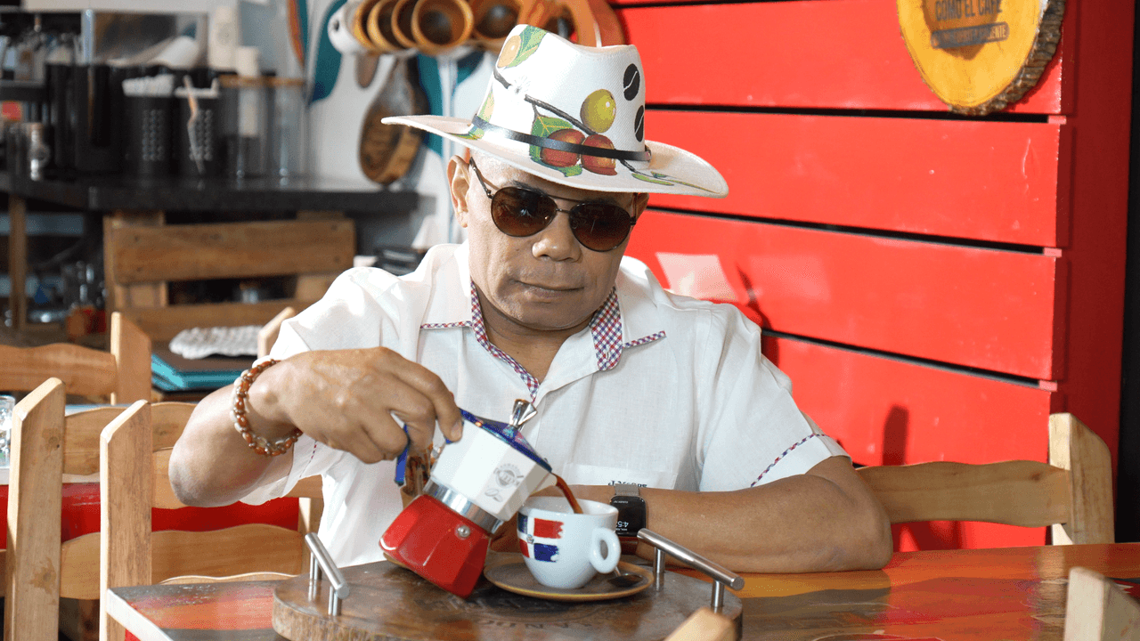Joe Veras sirve café de una greca en una cafetería, con sombrero y gafas oscuras